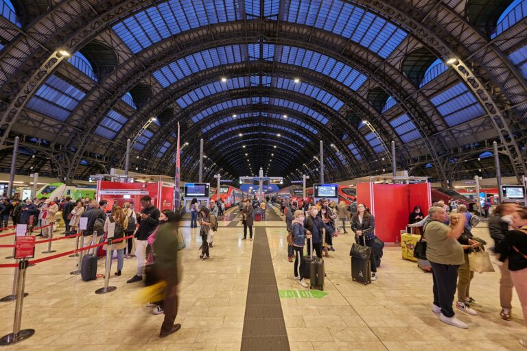 Travellers with suitcases on the platform concourse under the arched glass roof of Milano Centrale station in Italy, with trains at the platforms