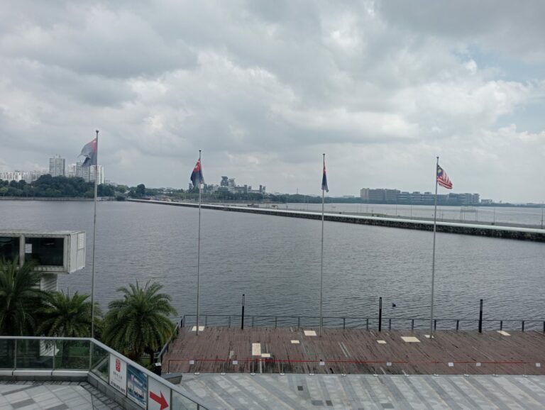 Johor-Singapore Causeway seen across the water from the Malaysian side, with flags on poles in the foreground