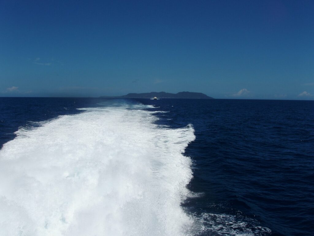 The white wake of a ferry leaving Green Island off Taitung, Taiwan, with the island on the horizon under a blue sky